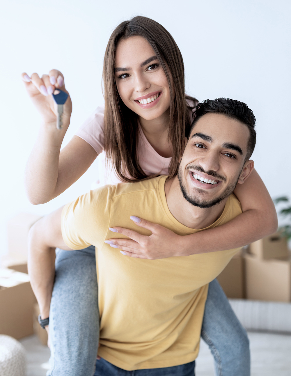 Cheerful Caucasian lady getting piggyback ride from her Arab boyfriend, showing house key indoors. Lovely multinational couple posing in new apartment on moving day